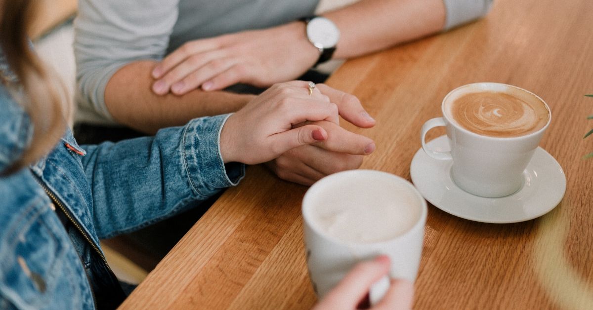 photo of a woman and man sitting at a table drinking coffee with the woman's hand on top of his