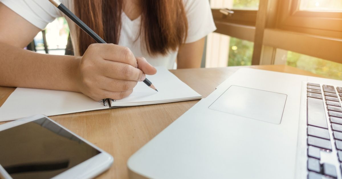 photo depicts a girl writing with a computer and phone at her desk