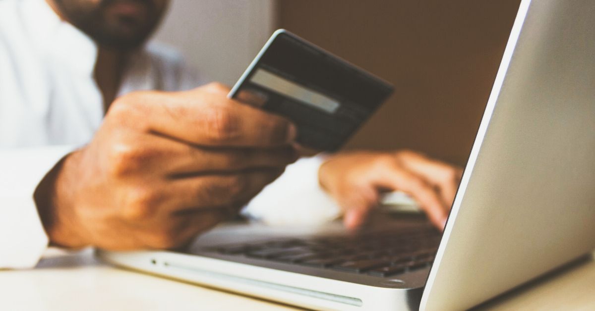 close up photo of a man's hand holding a credit card as he types on his computer