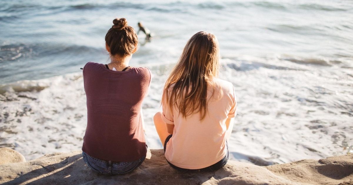 two young women sitting side by side on beach facing away from camera