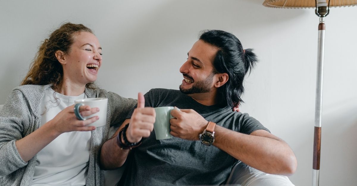 a couple holding mugs sitting on the couch together laughing