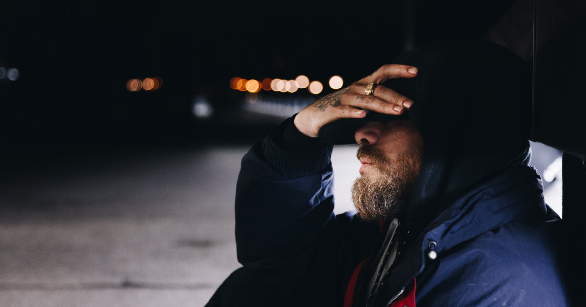 unshaven man sitting on dark street with head in hand