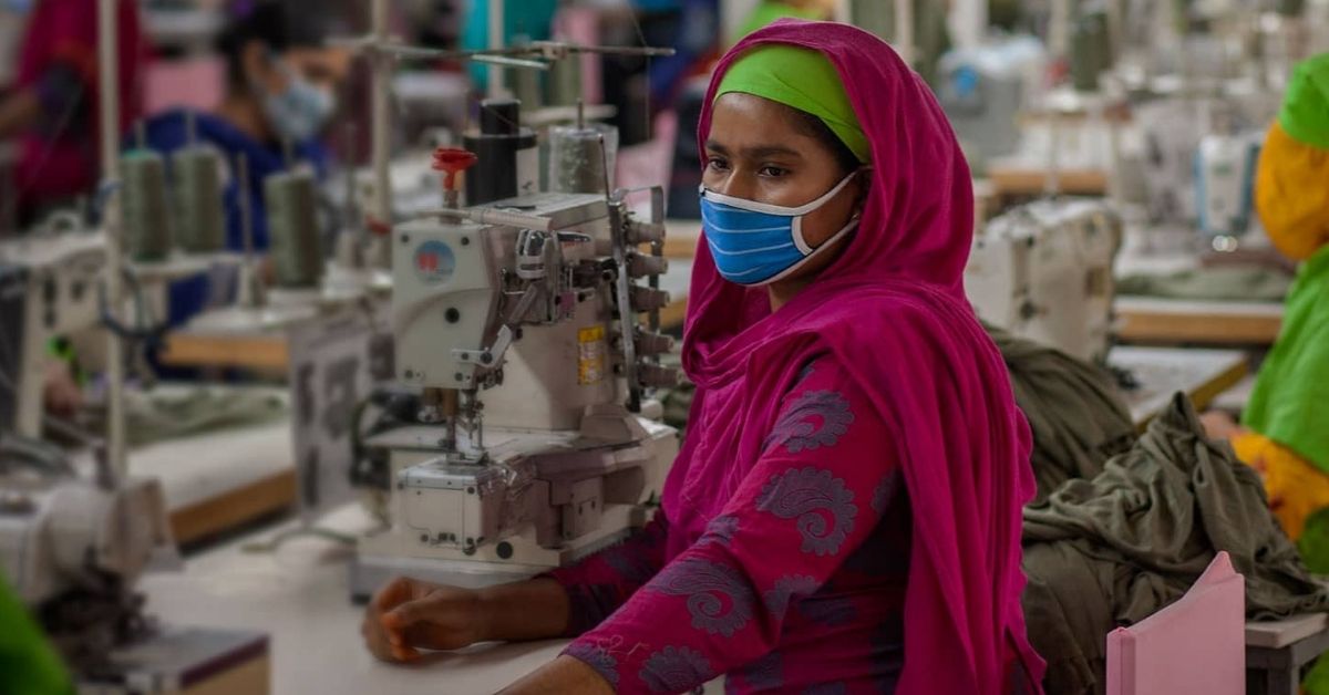 a female worker stands beside a sewing machine wearing a mask