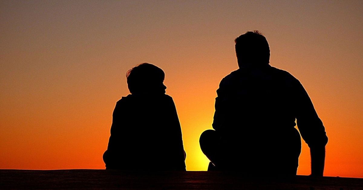 the silhouette of a father and son sitting on the ground talking from behind