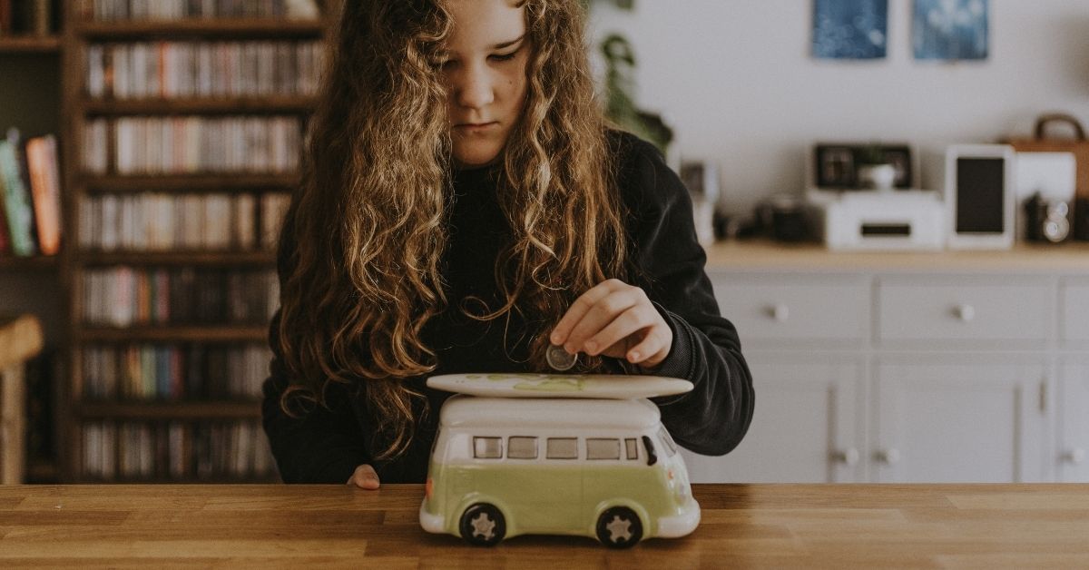 a young girl puts a dollar into a kombi van shaped piggy bank